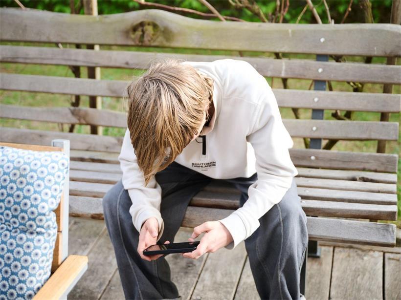 Ein Junge guckt auf sein Smartphone, während er auf einer Bank in einem Garten sitzt