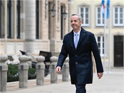 Georges Mischo steht vor dem historischen Parlamentsgebäude bei klarem Himmel, symbolisch für Politik und Demokratie.