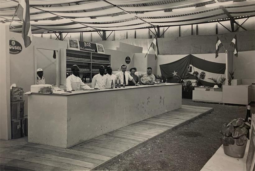Luxemburgischer Stand auf der Messe in Léopoldville, dem heutigen Kinshasa, im Jahr 1951, im Hintergrund die Flagge Luxemburgs neben jener des Belgisch‑Kongo sowie ein Porträt der Großherzogin Charlotte in der Mitte
